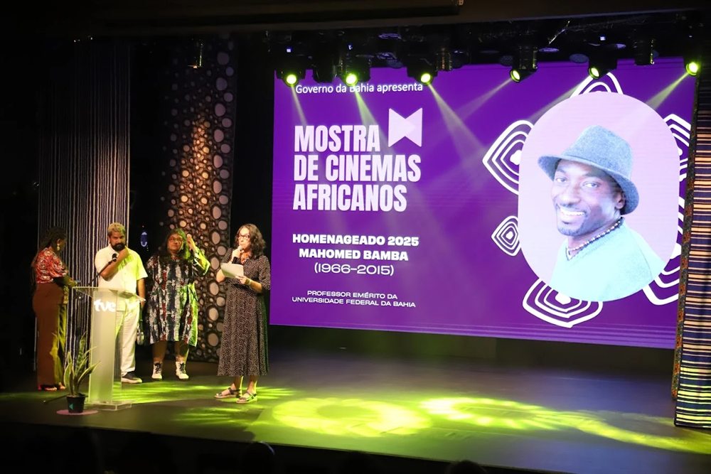 Homenagem a Mahomed Bamba na Mostra de Cinemas Africanos 2025: no palco, organizadores diante do telão com a foto do homenageado